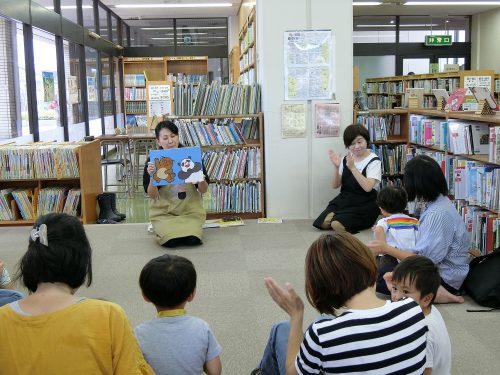 図書館の一角で床に座り絵本の読み聞かせをする女性とそれを聞く3～4組の親子の様子