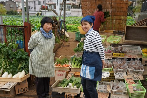直売所に陳列した野菜の前で微笑む女性の写真