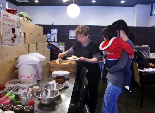 子どもを抱きかかえた女性に料理の盛りつけを手伝うメンバーの様子の写真