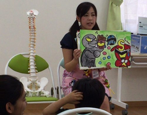 背骨の骨格標本を横に置き、子ども達に手作りの紙芝居を披露する女性の写真
